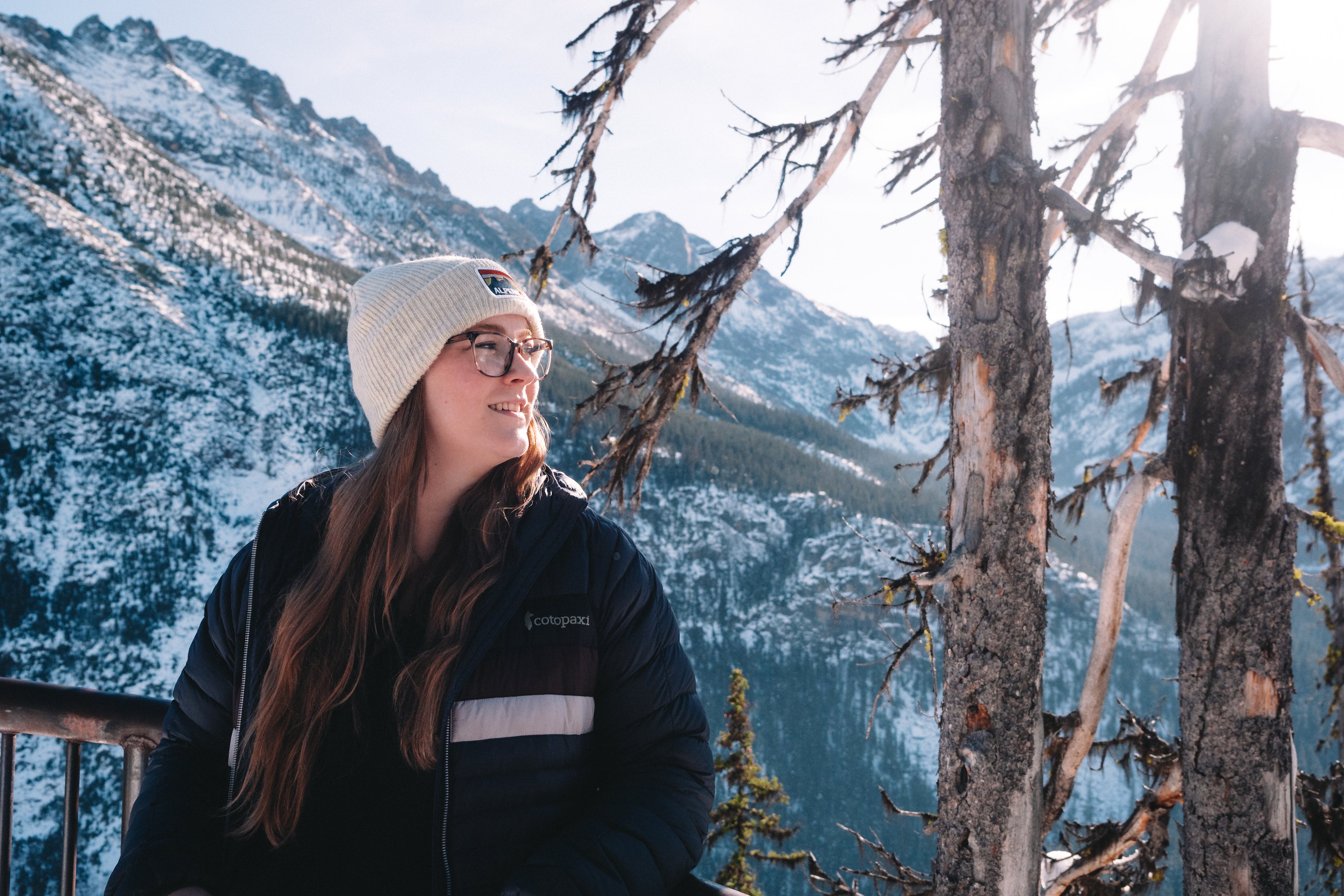 Young woman wearing a cream-colored beanie with Alpenglow logo, standing outdoors against a snowy mountain landscape.