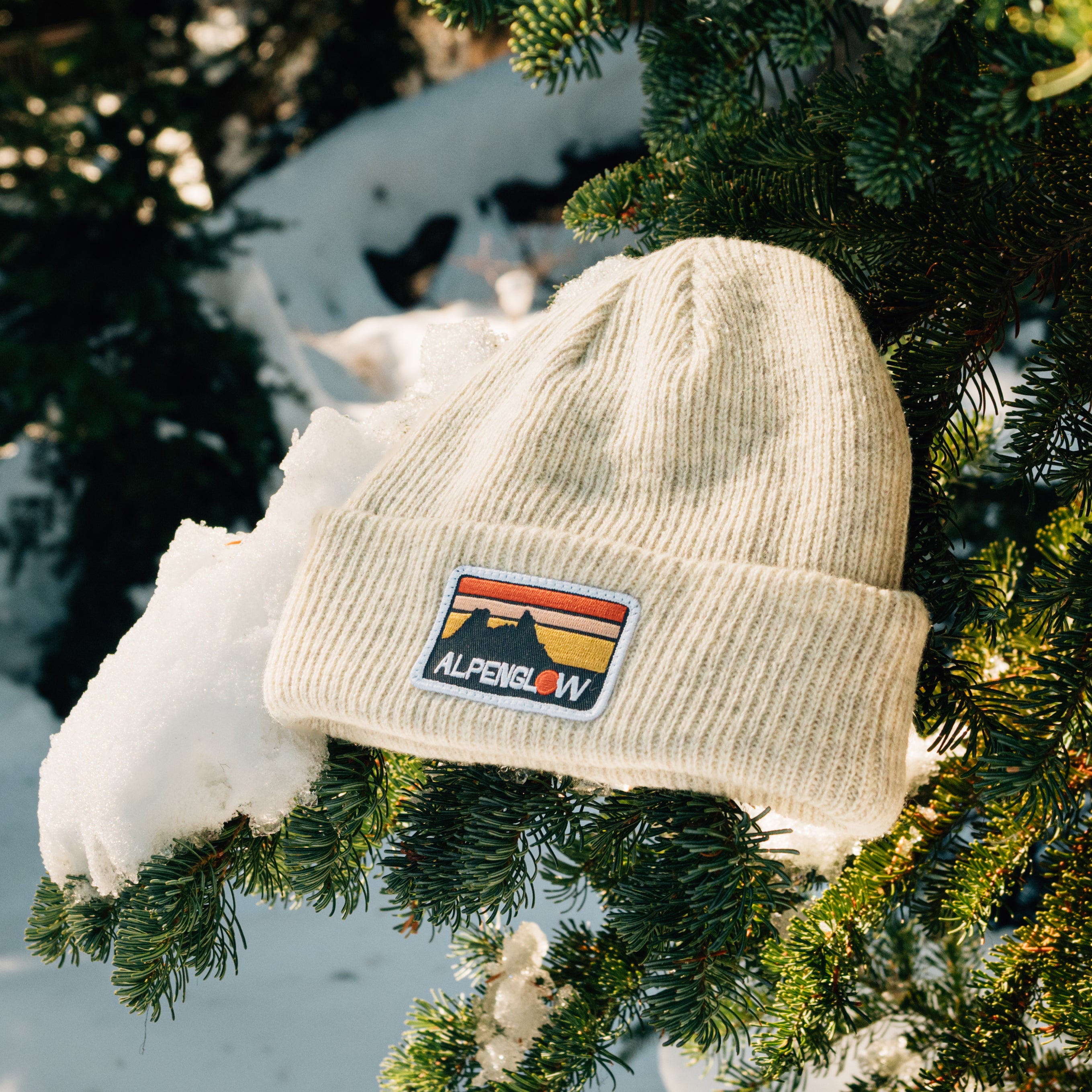 A beige knit hat with a colorful "Alpenglow" patch rests on a snow-covered evergreen branch.